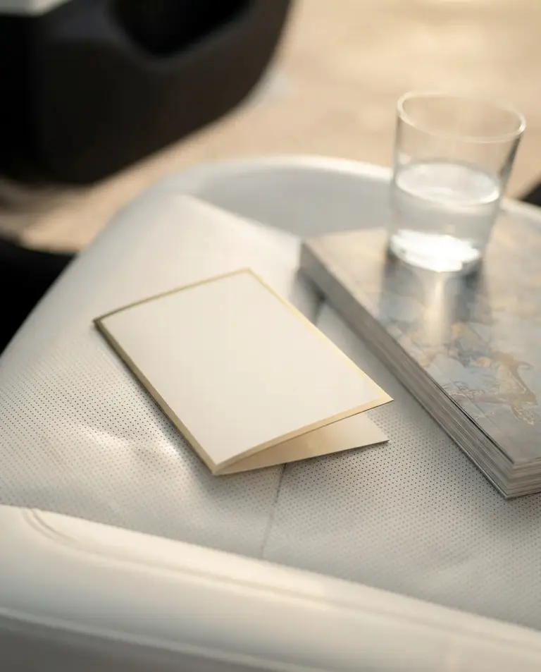 Carte ivoire à liseré laiton posée sur le cuir blanc de la banquette, à côté d’un magazine et d’un verre d’eau